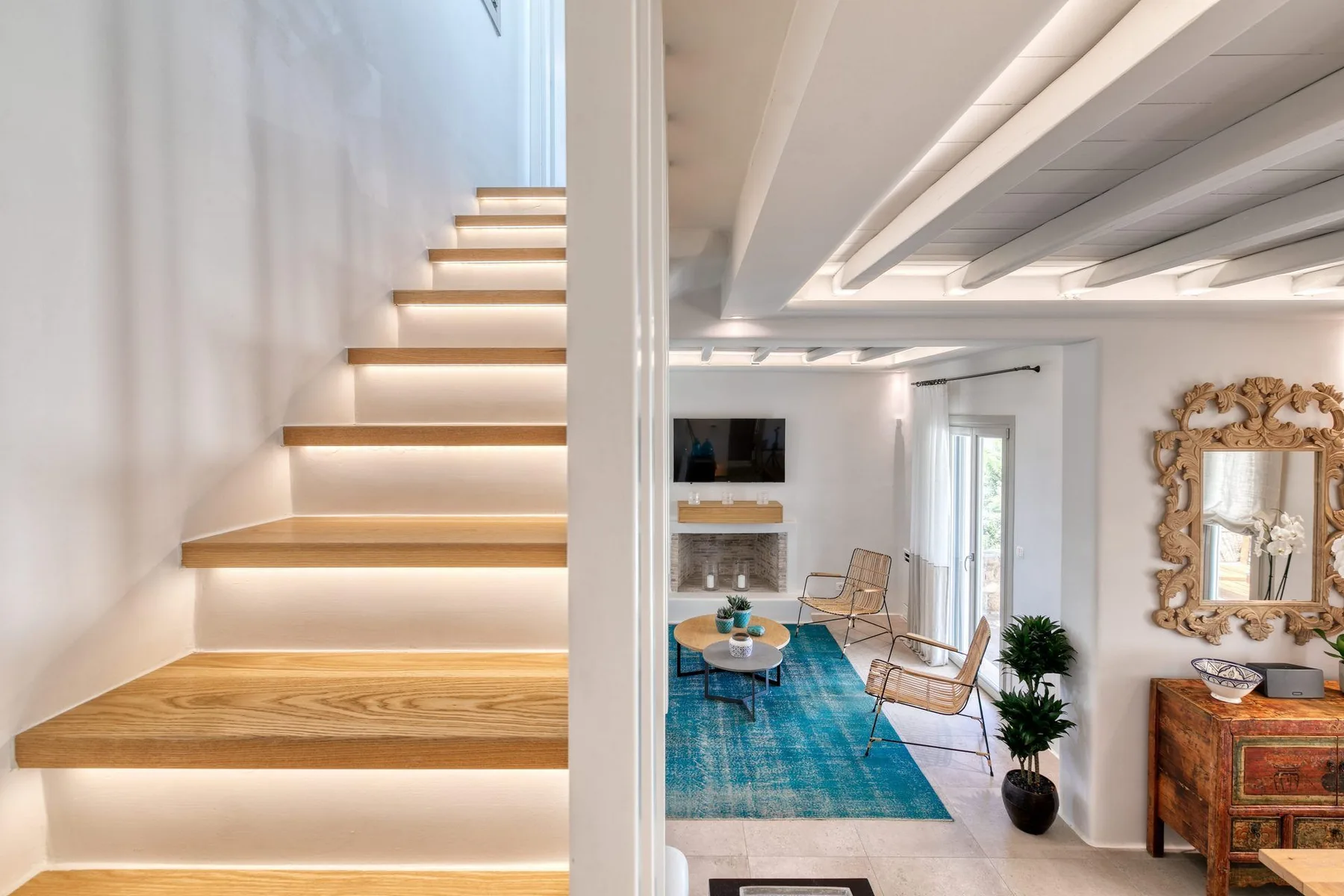 A bright living room with a blue rug, seating, and an illuminated staircase.