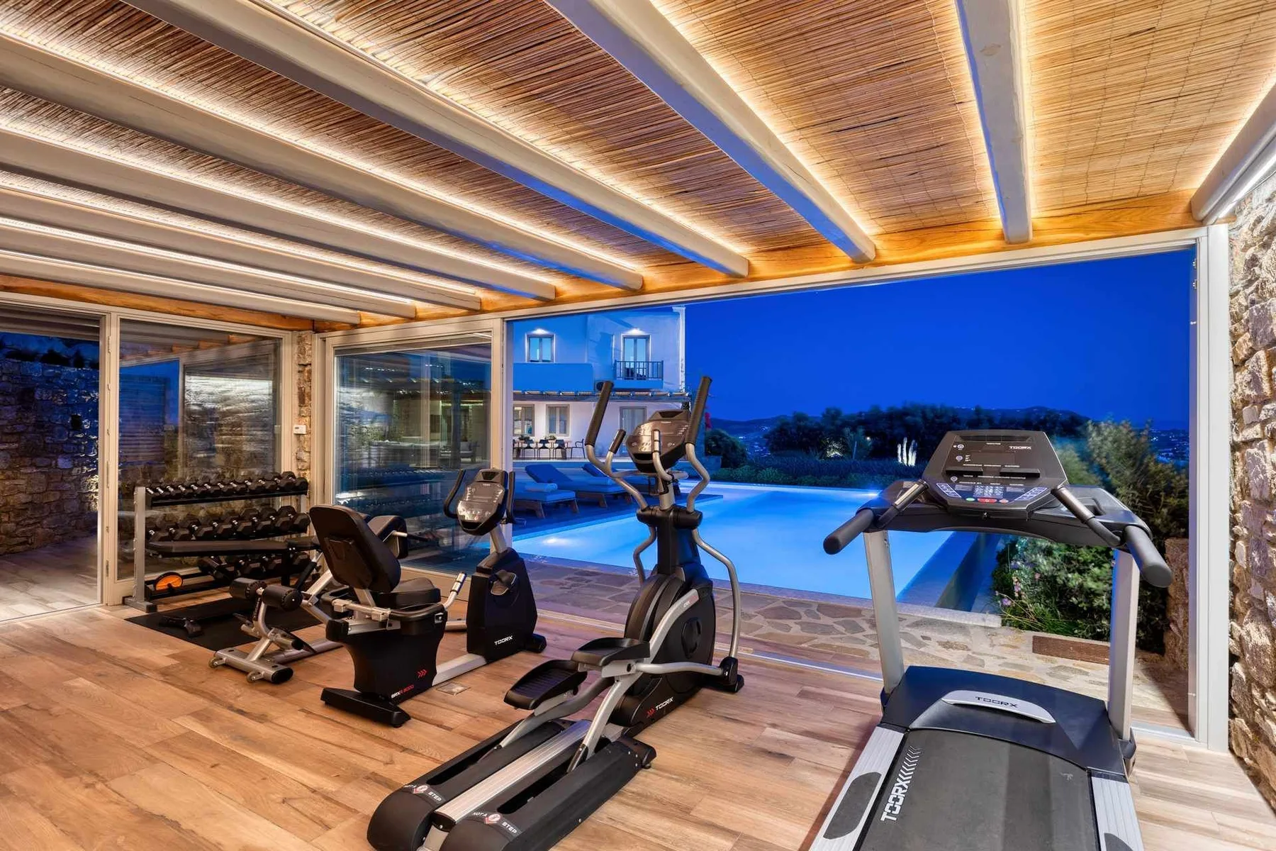 An indoor gym with exercise equipment overlooks a swimming pool at dusk.