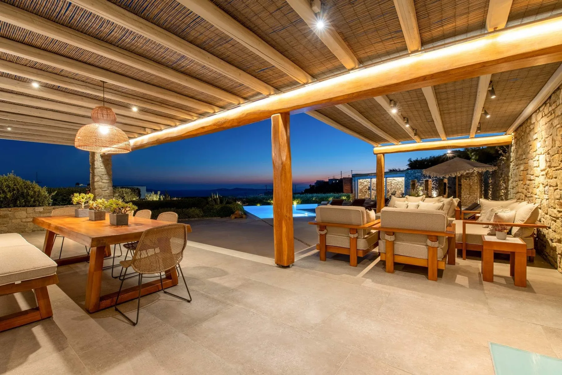 A covered outdoor dining and lounge area opens onto a pool and evening view.