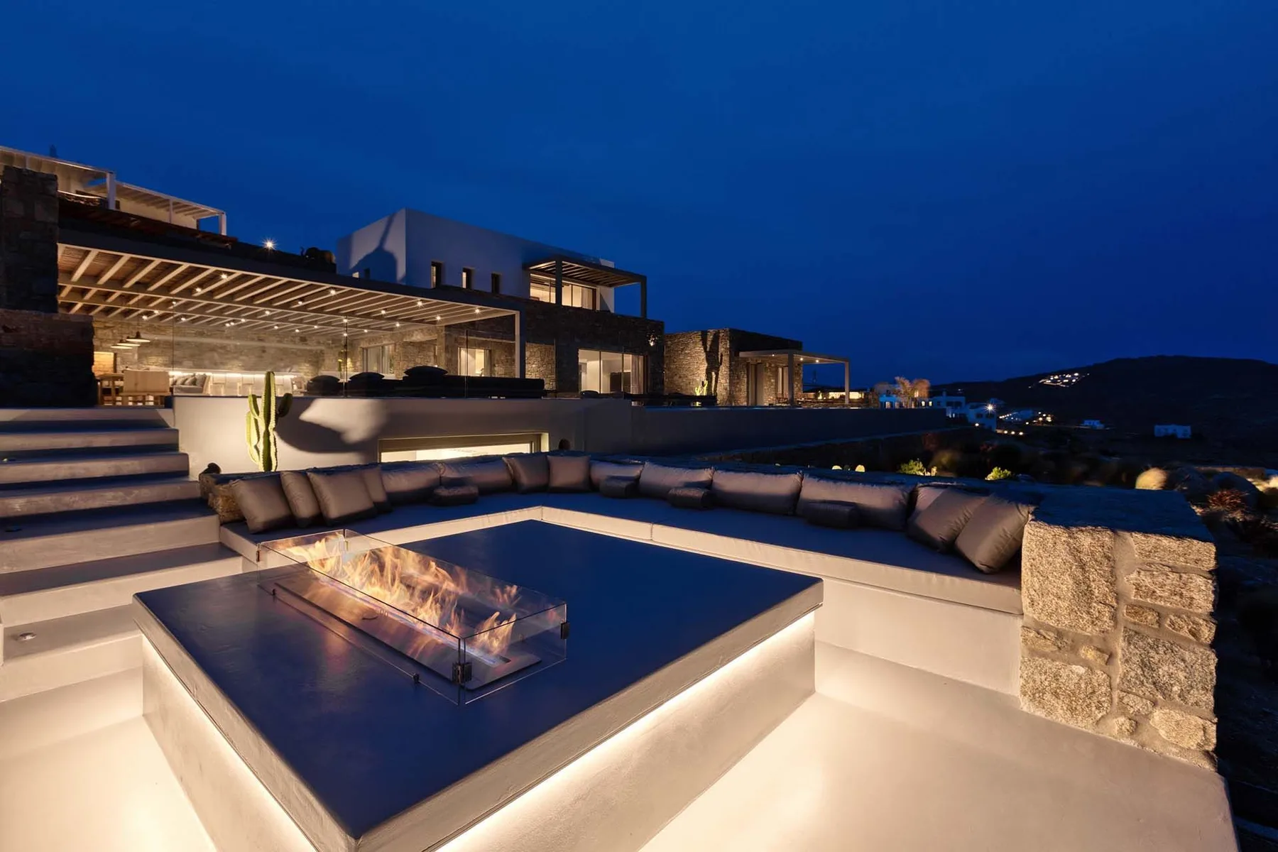 Una zona de estar exterior iluminada, con asientos integrados y una chimenea central al anochecer.