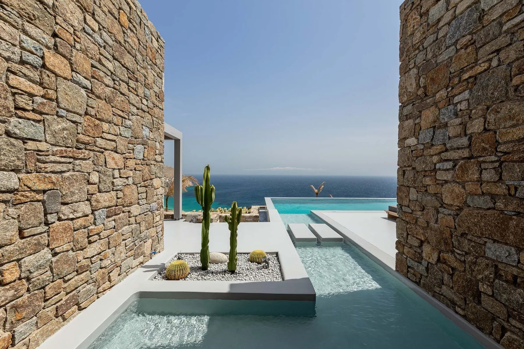 Una terraza moderna con piscina, enmarcada por muros de piedra y el mar al fondo.