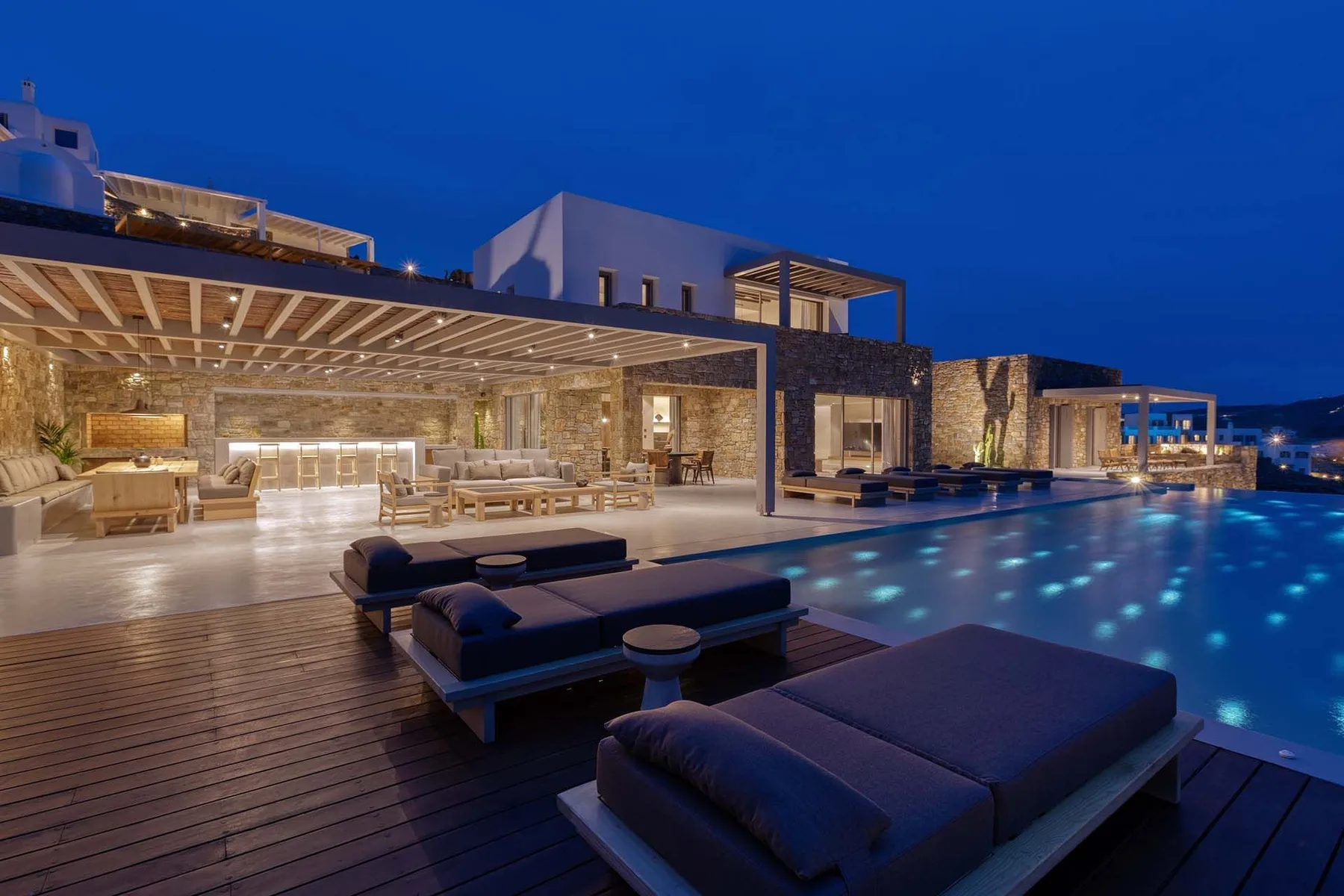 Una piscina exterior iluminada junto a un edificio moderno, con asientos en la terraza.