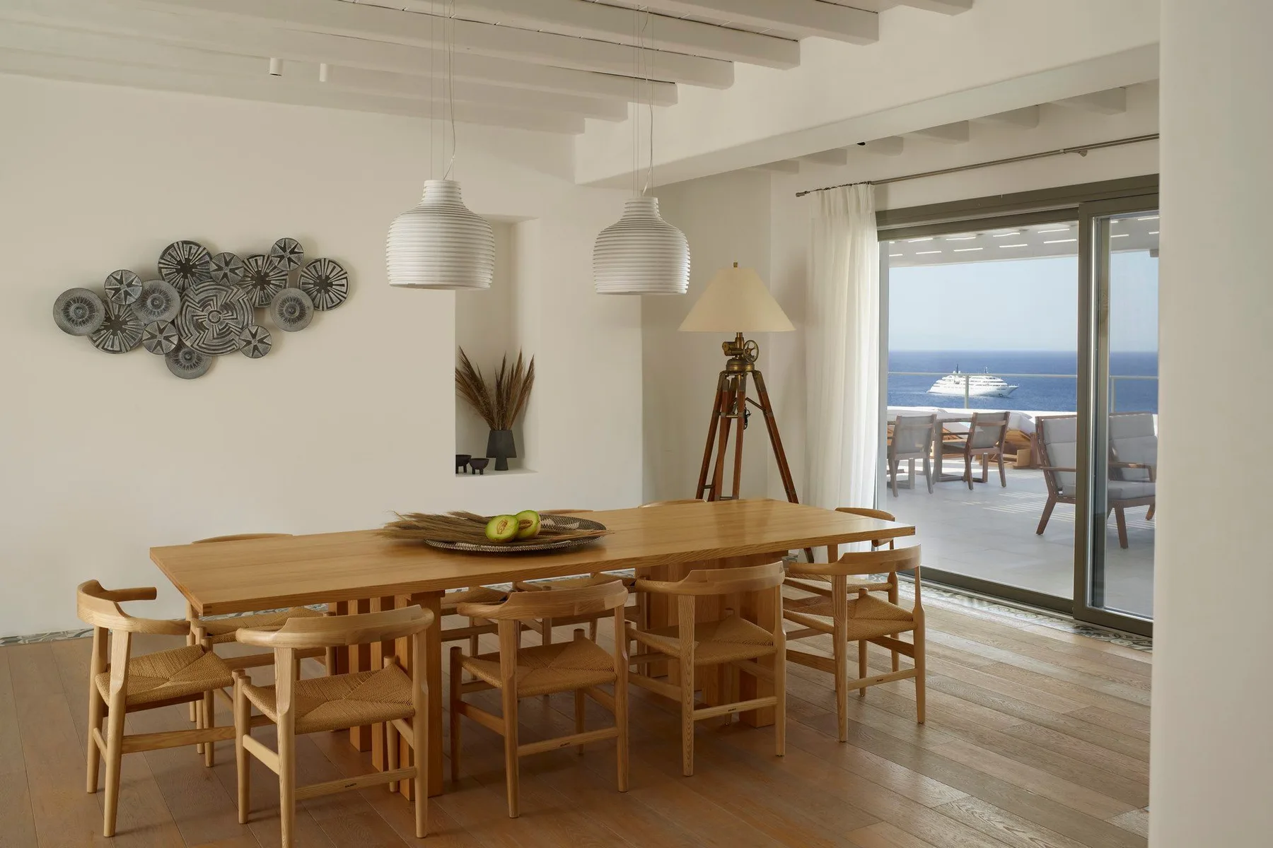 A bright dining area has a long wooden table beside sliding glass doors opening to a sea view.