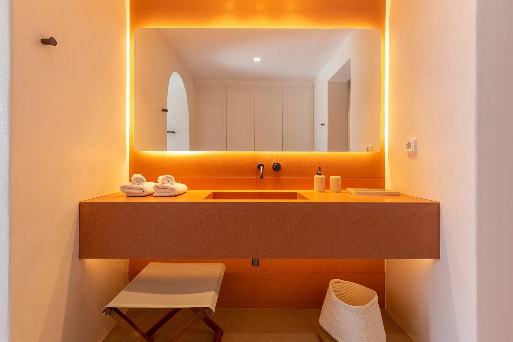A bathroom vanity with a wide mirror, warm backlighting, and a stool below.
