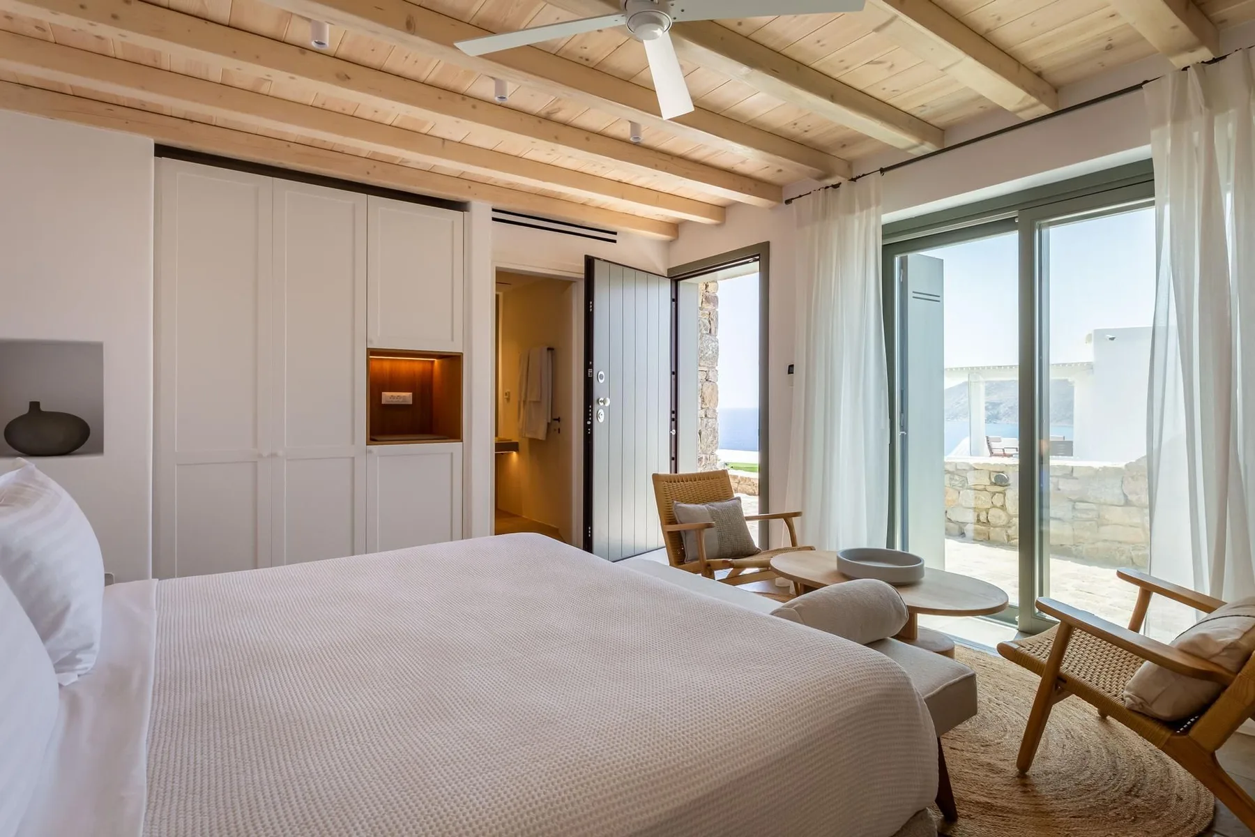 A bright bedroom with a large bed and a sitting area beside glass doors.