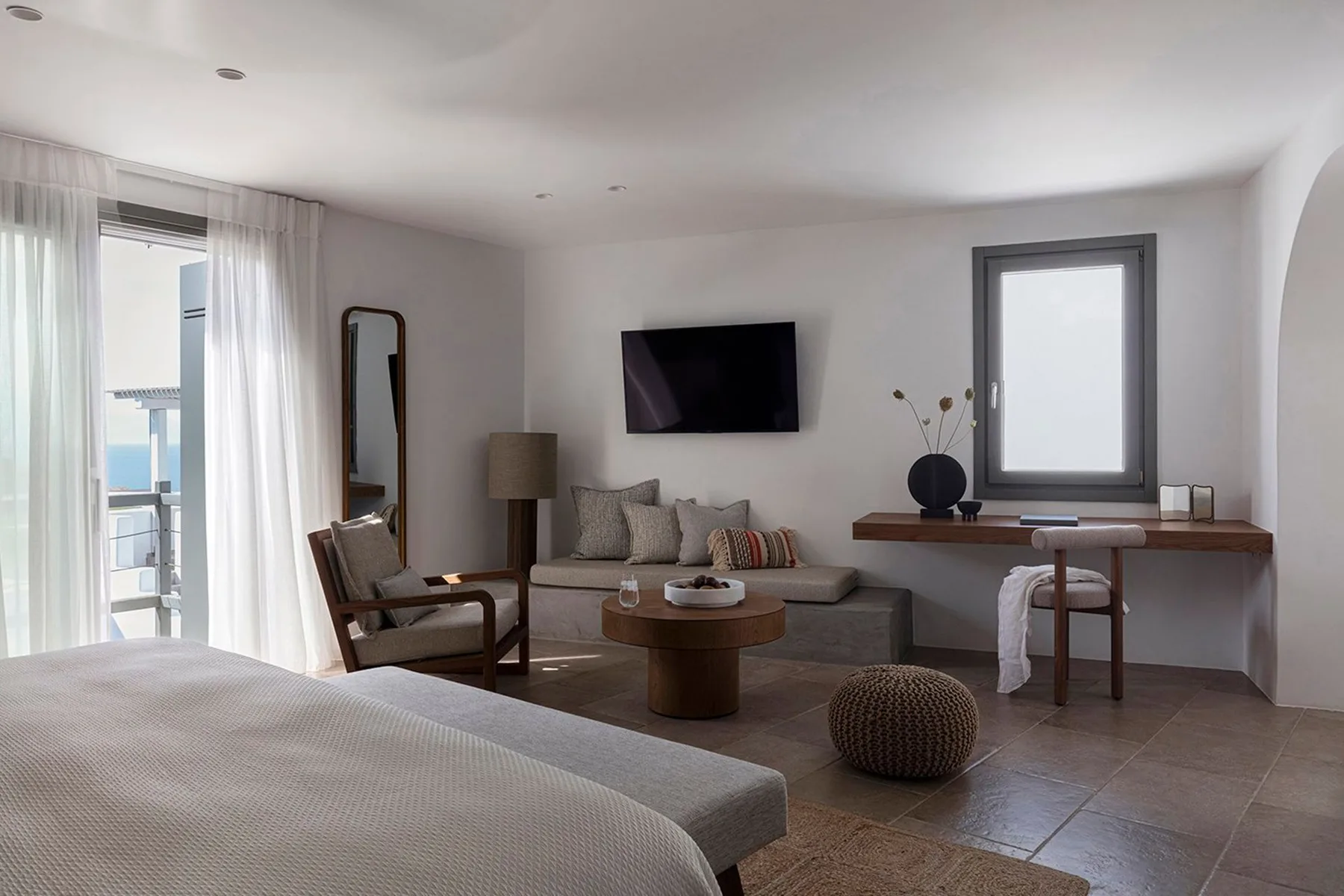 A bright bedroom with a bed, seating area, and desk beneath a window.