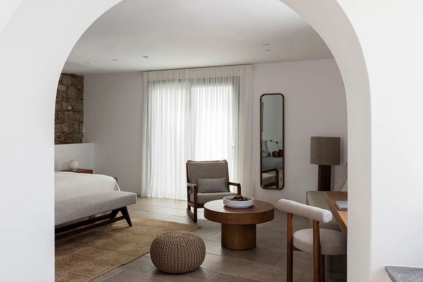 A bedroom sitting area with a bed, armchair, and large curtained window.