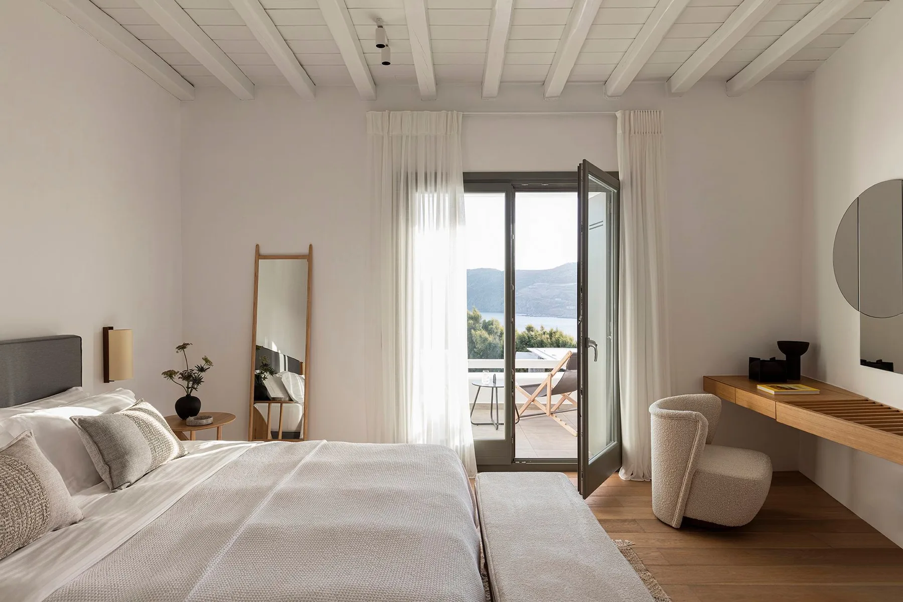 A bright bedroom with a bed, a desk, and an open glass door to a balcony.