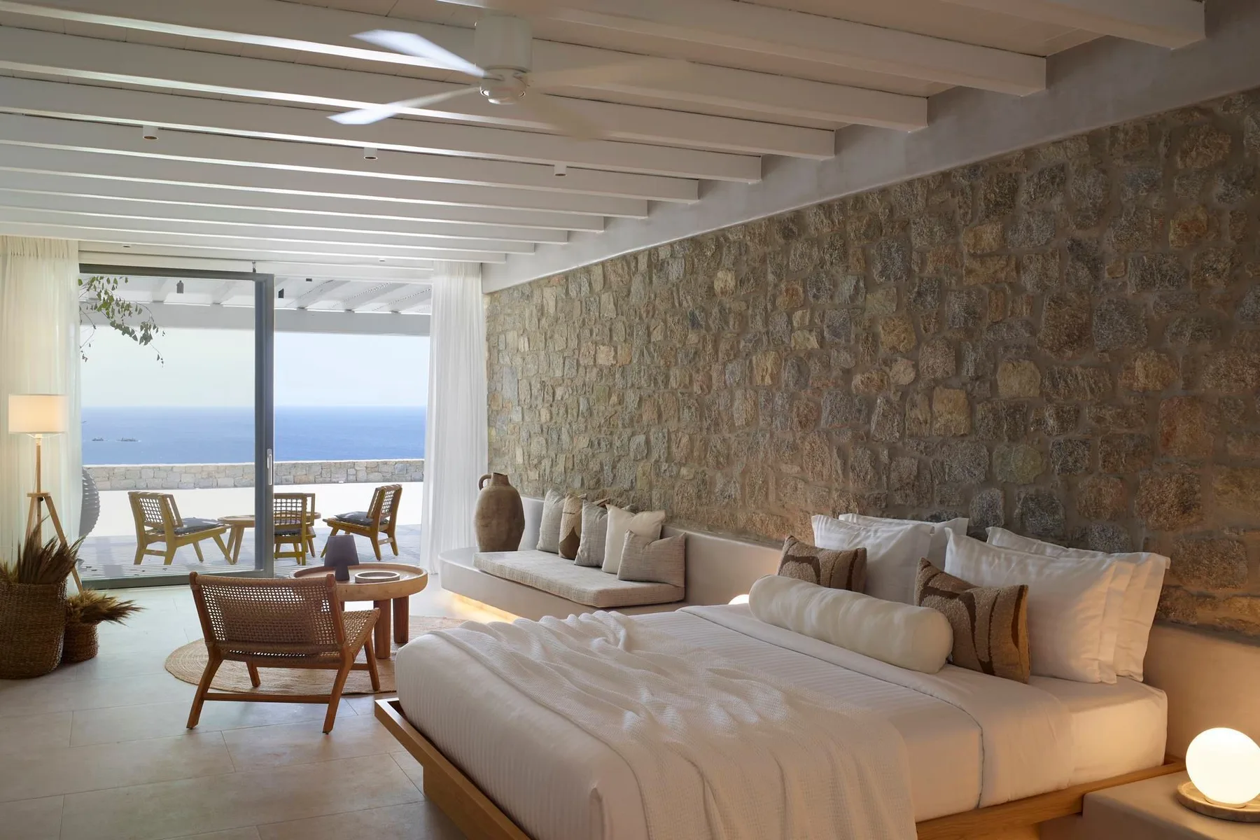 A bedroom with a large bed, seating, and a view of the sea through glass doors.