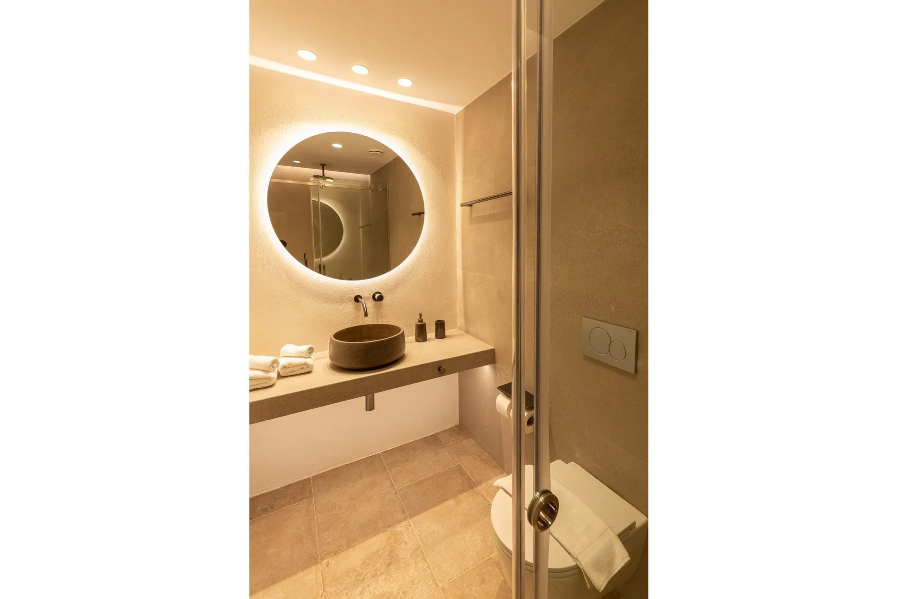 A warm-lit bathroom with a round mirror above a sink.