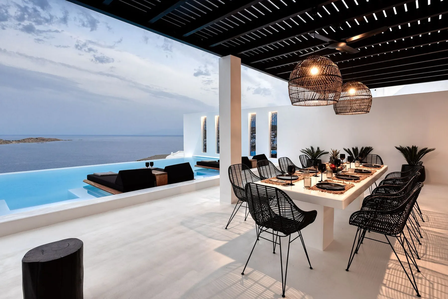 An open-air dining area with a long table beside a pool overlooking the sea.