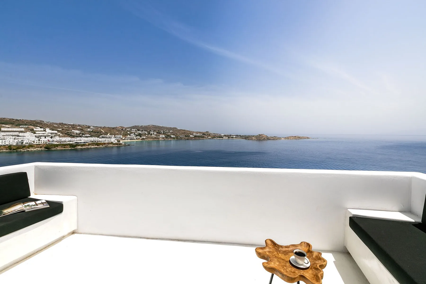 A white terrace overlooks the sea, with dark cushioned seating and a small table.