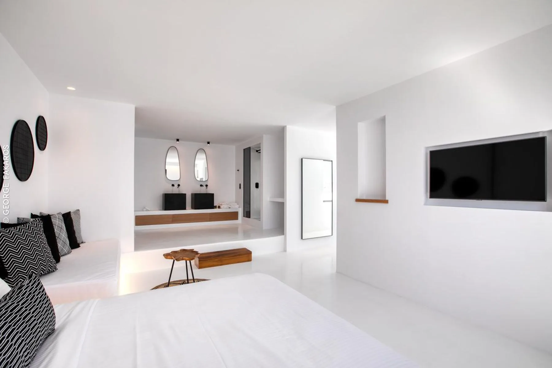 A bright bedroom with white bedding, patterned cushions, and a television.