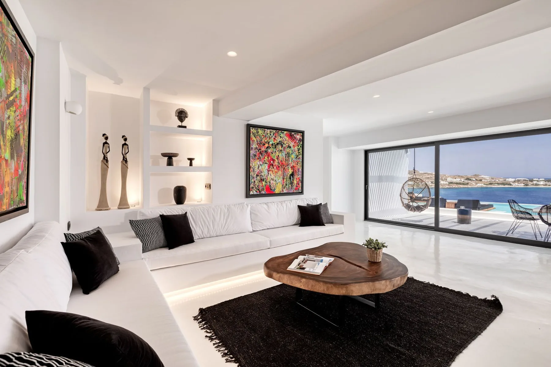 A bright living room has white seating, colorful artwork, and a large window overlooking the water.