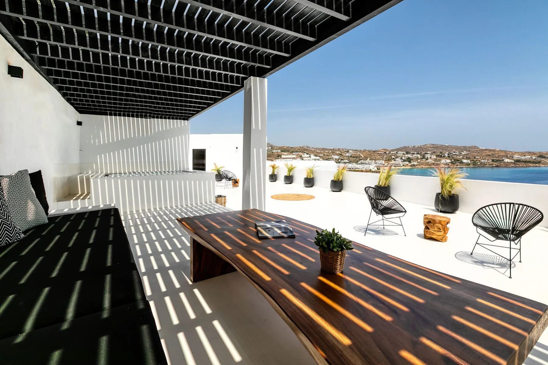 A rooftop terrace has a long outdoor table, seating, and potted plants.