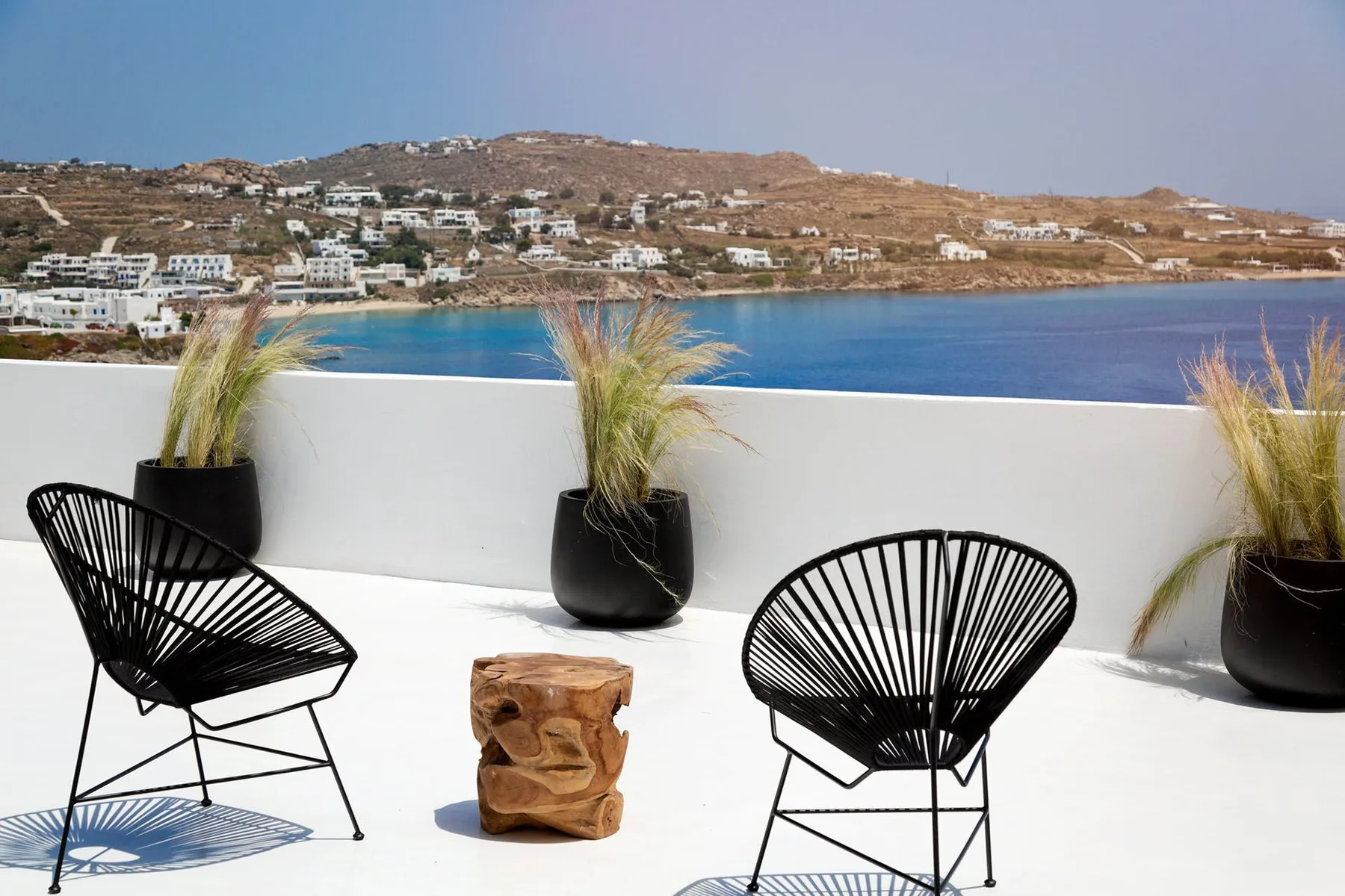 A sunny seaside terrace with black seating and potted grasses overlooking blue water.