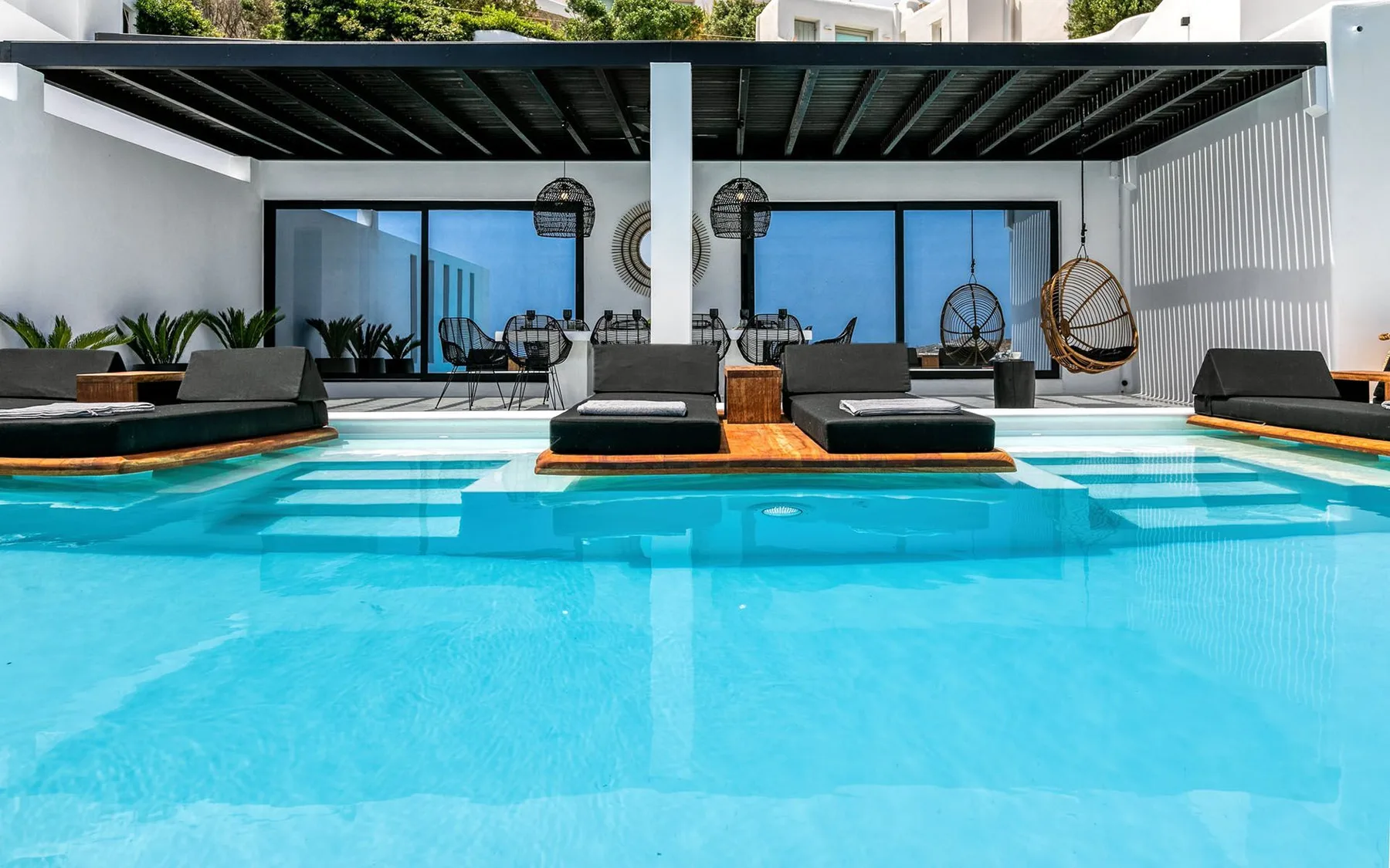 A swimming pool sits beside a shaded seating area with large glass doors.