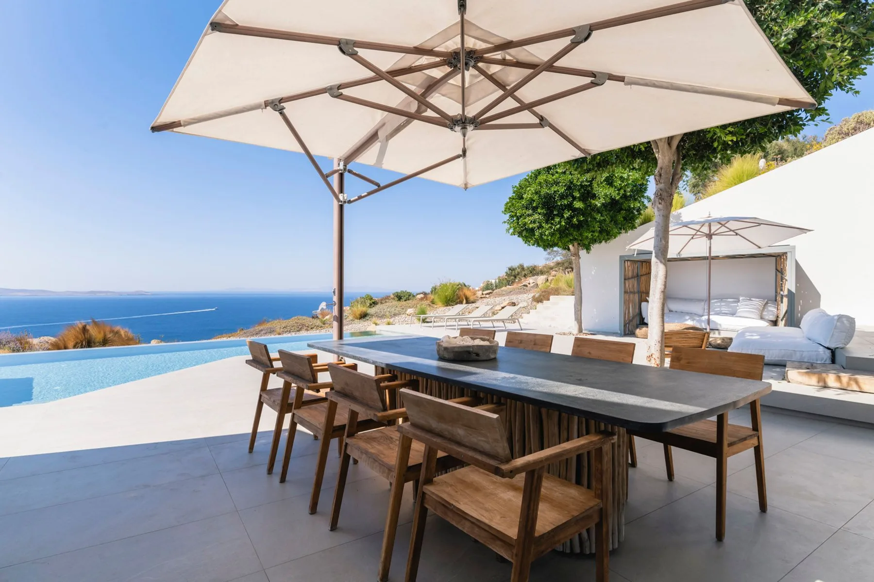 An outdoor dining table under a large umbrella overlooks a pool and the sea.