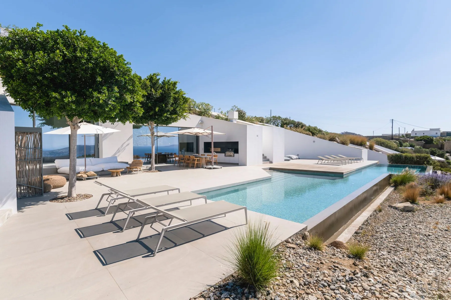 A modern poolside patio with lounge seating, trees, and a white building.