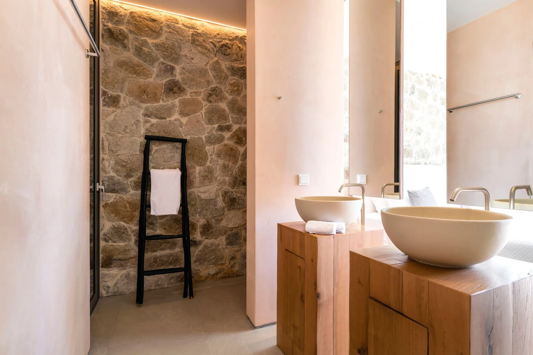 A bright bathroom with two vessel sinks on wooden vanities and a stone wall.