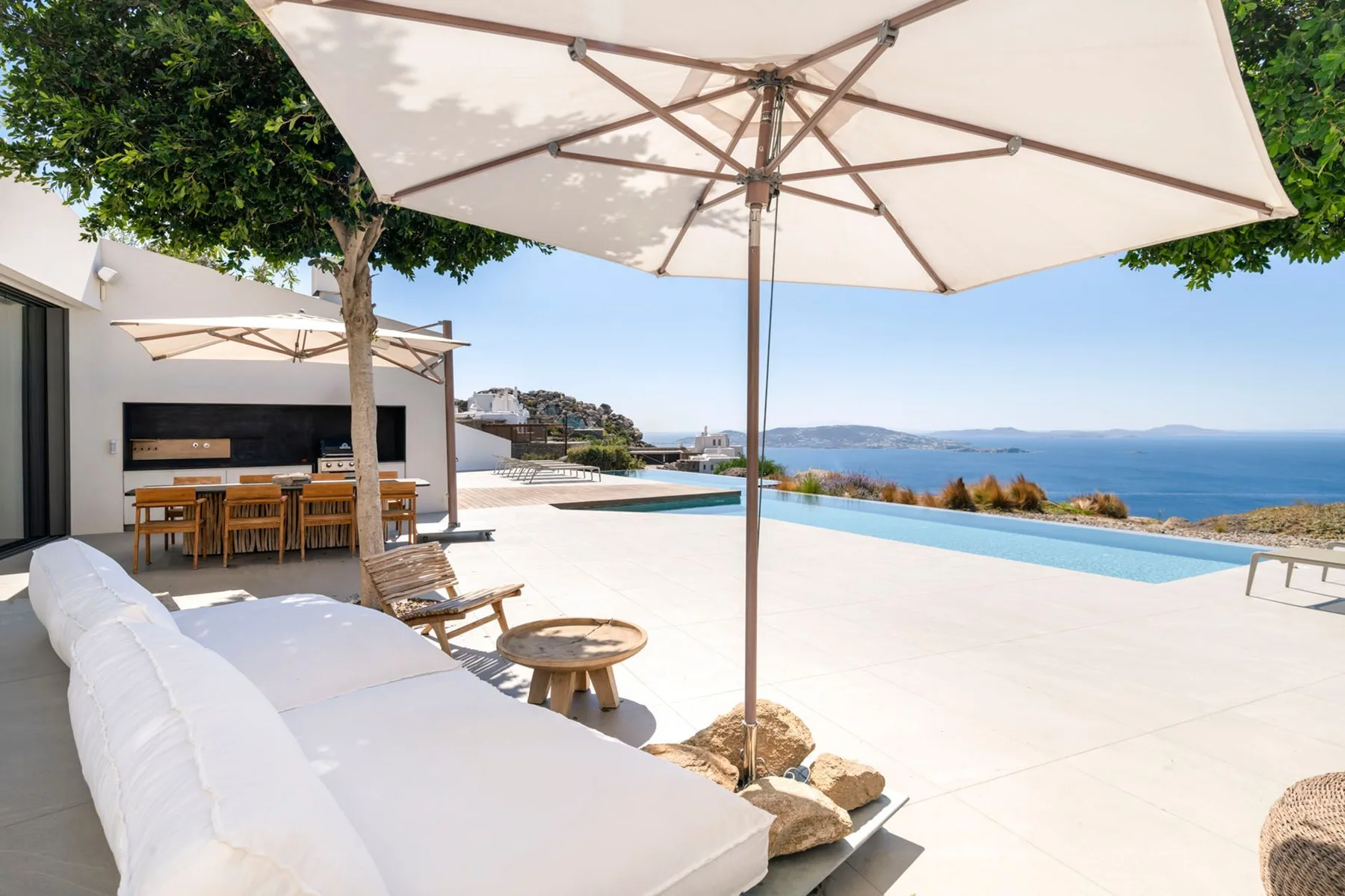 An outdoor pool terrace overlooks the sea, with shaded seating beneath a large umbrella.
