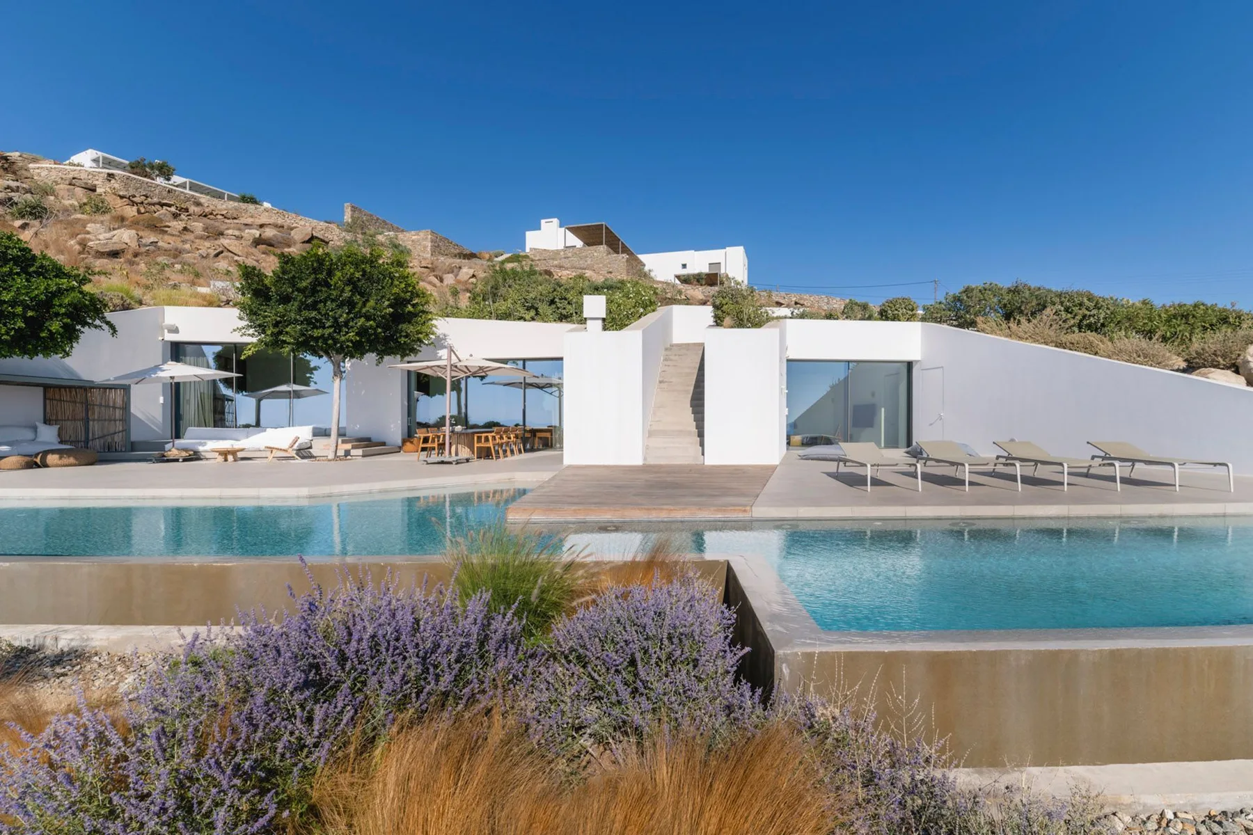 A modern white building overlooks a swimming pool and landscaped hillside.
