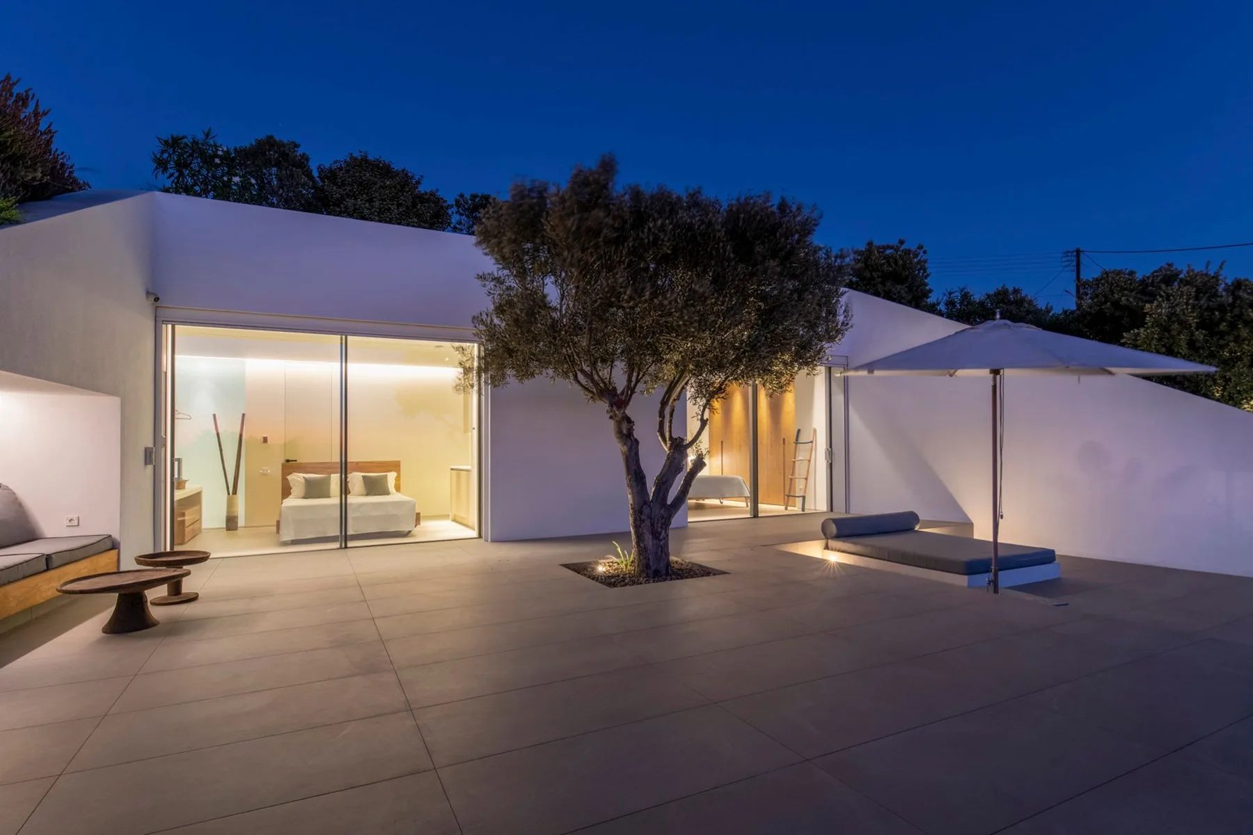A modern home opens onto a paved patio with a tree and umbrella.