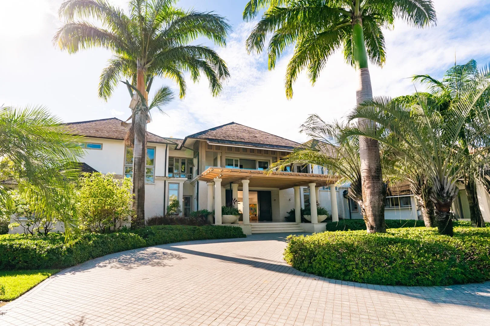 A large house stands behind a paved driveway, surrounded by palm trees and greenery.