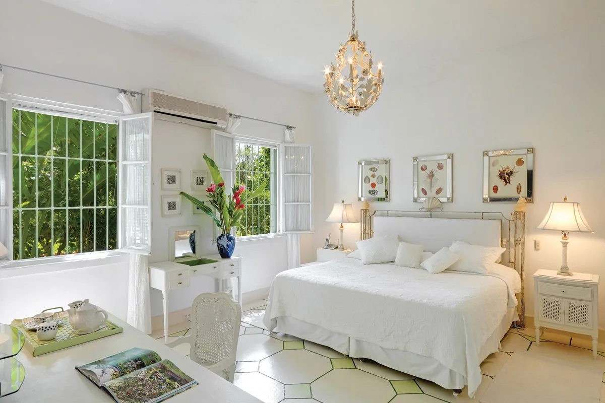 Un dormitorio luminoso con una cama blanca, grandes ventanas y una lámpara de araña.