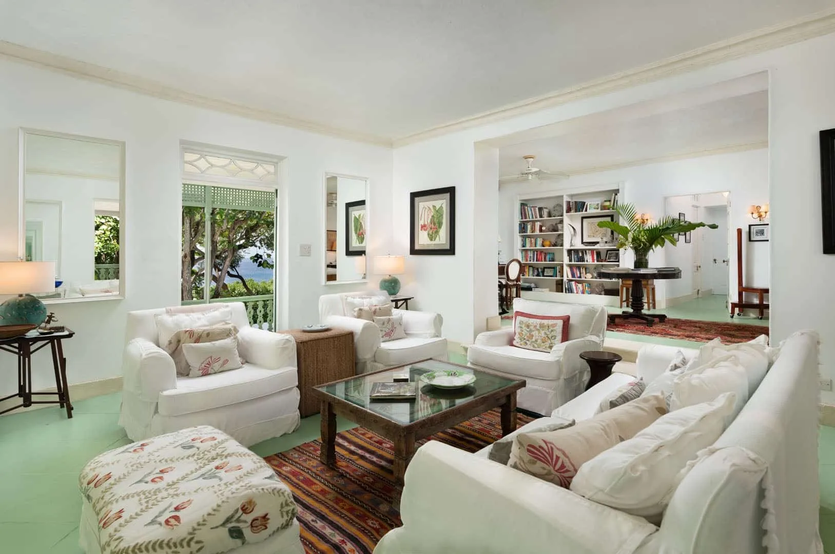 A bright living room with white seating around a glass coffee table and bookshelves beyond.
