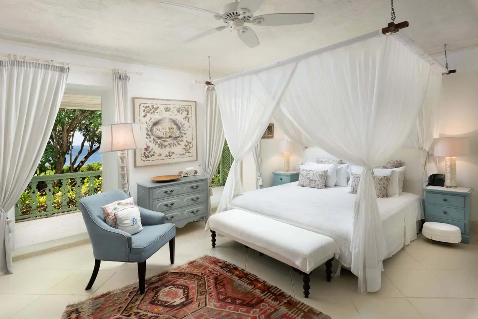 A bright bedroom with a curtained bed, seating, and a leafy balcony view.