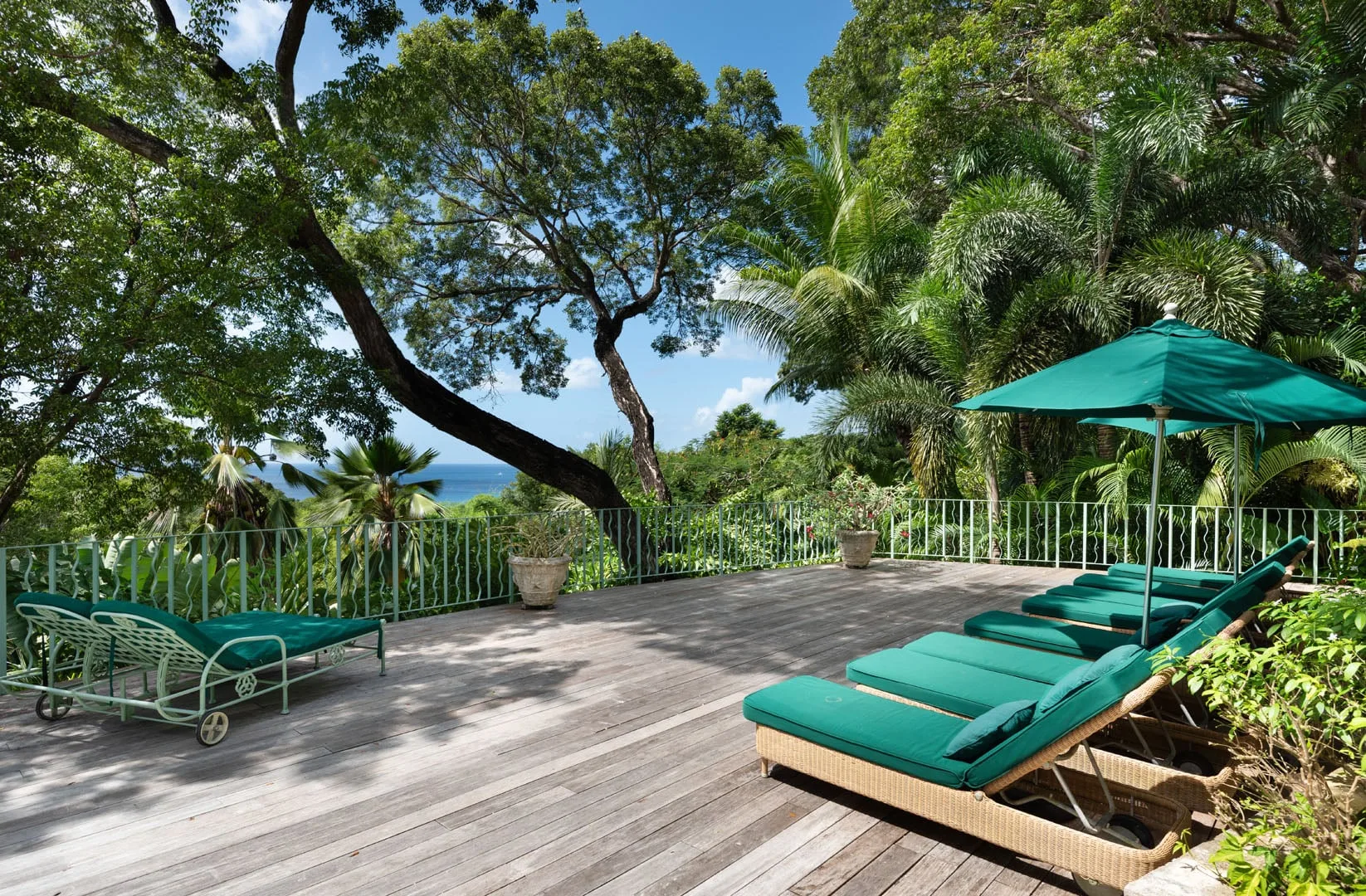 An outdoor deck has teal lounge seating beneath leafy trees and a matching umbrella.