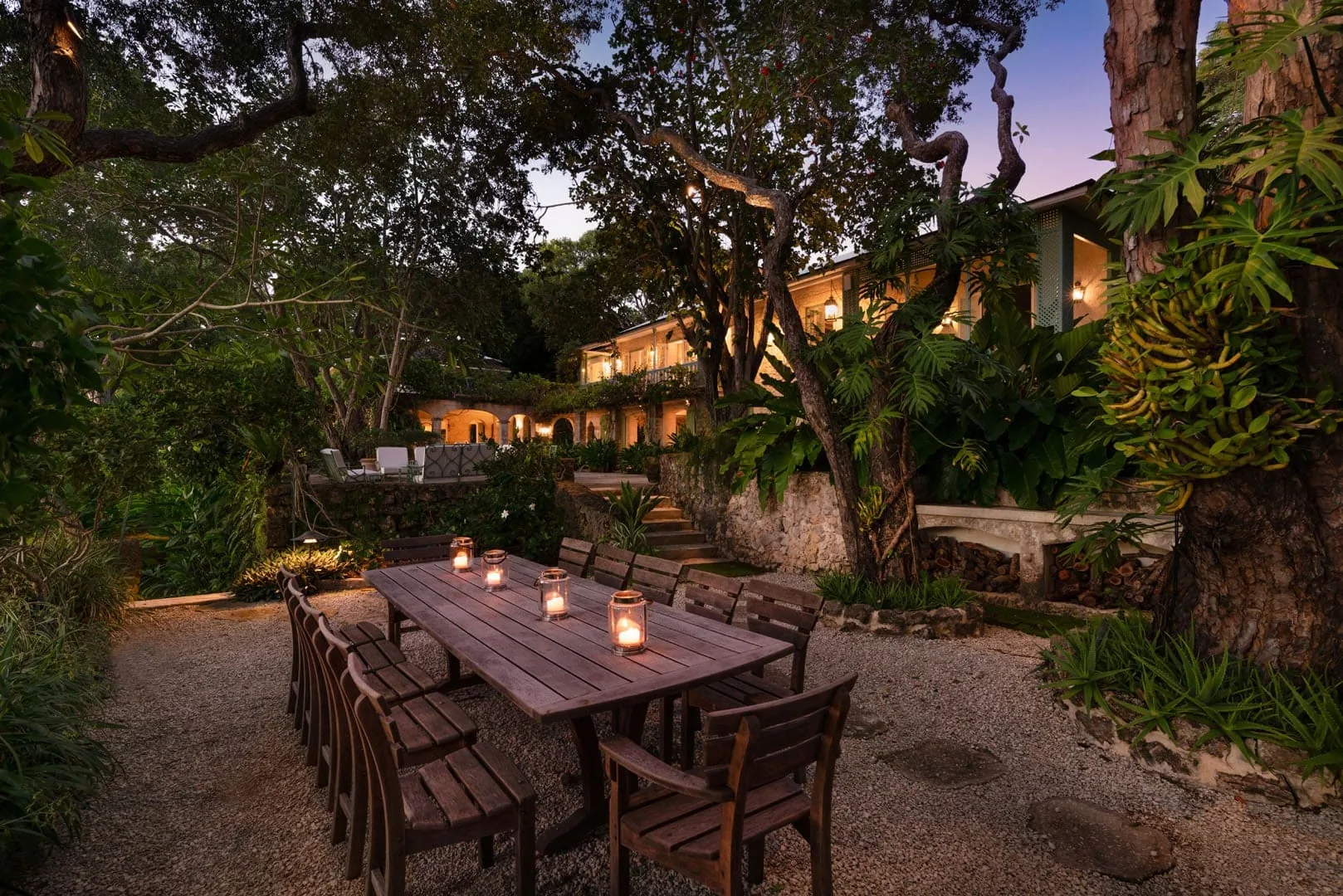 An outdoor dining area with a long wooden table surrounded by trees and greenery.