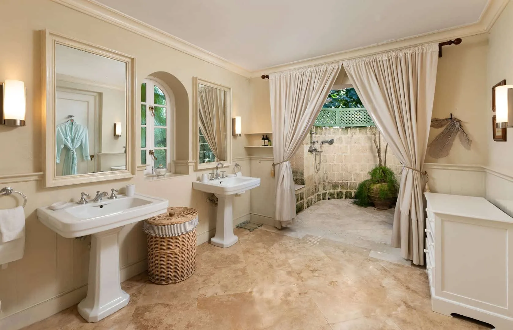 A bright bathroom with two sinks and curtains opening onto a small garden.