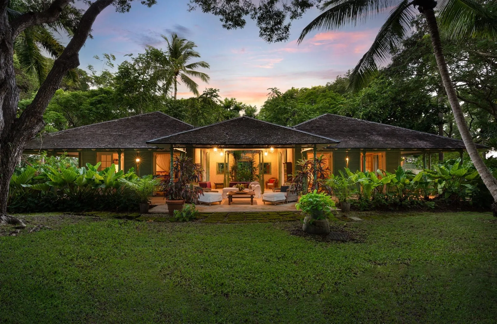 A single-story building with a covered patio sits amid lush tropical greenery at dusk.