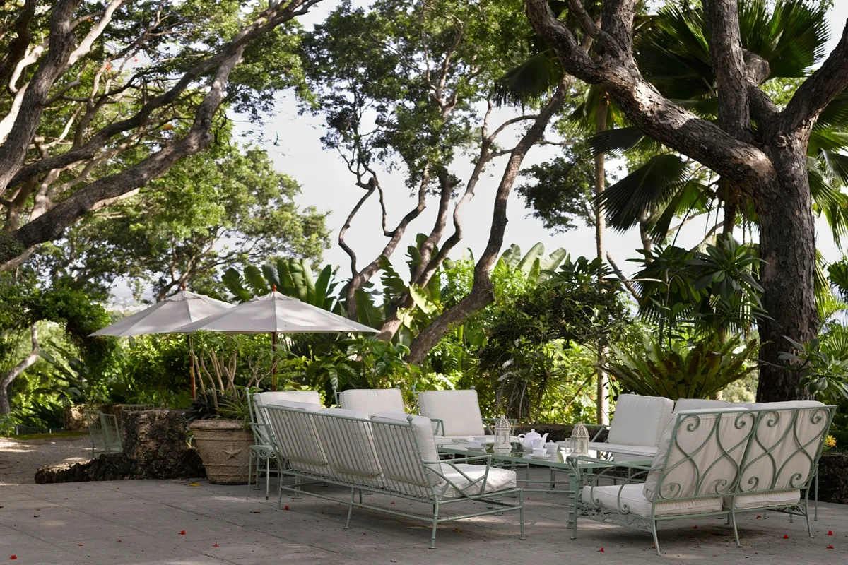 An outdoor seating area sits beneath leafy trees beside a patio umbrella.