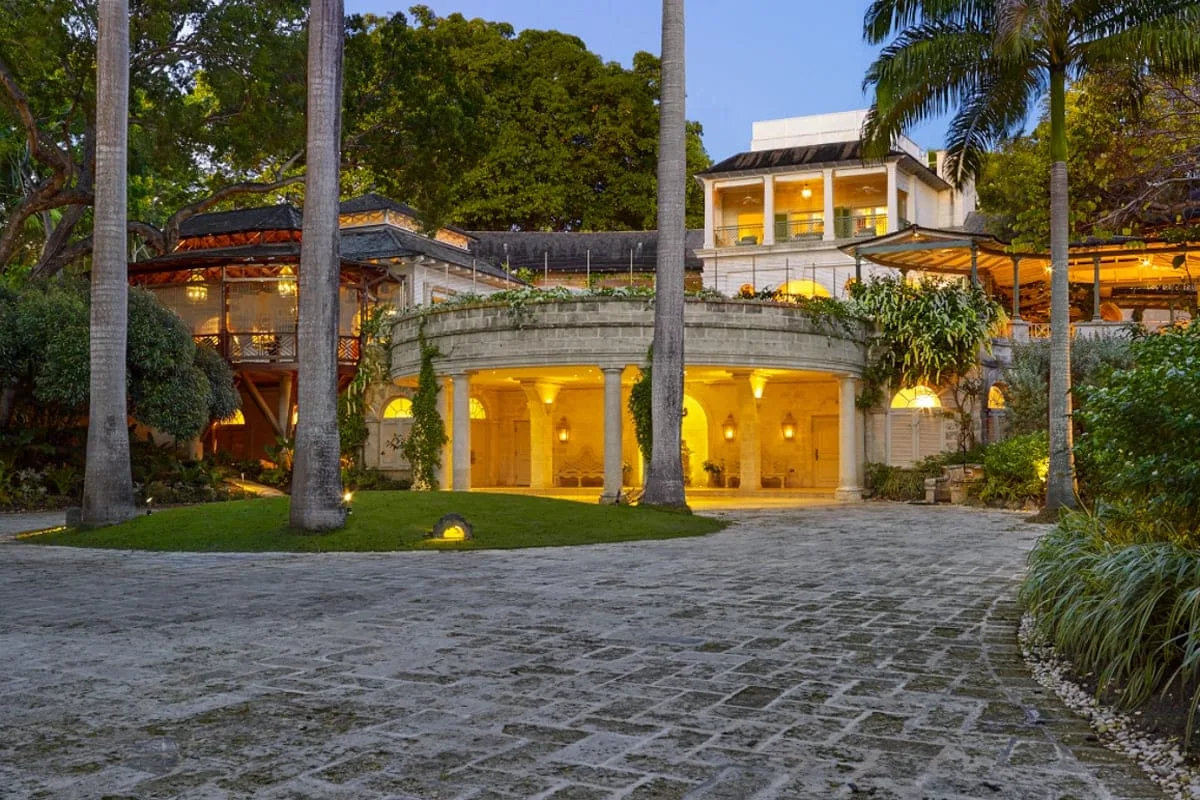Un edificio iluminado se alza detrás de una entrada pavimentada y vegetación tropical.
