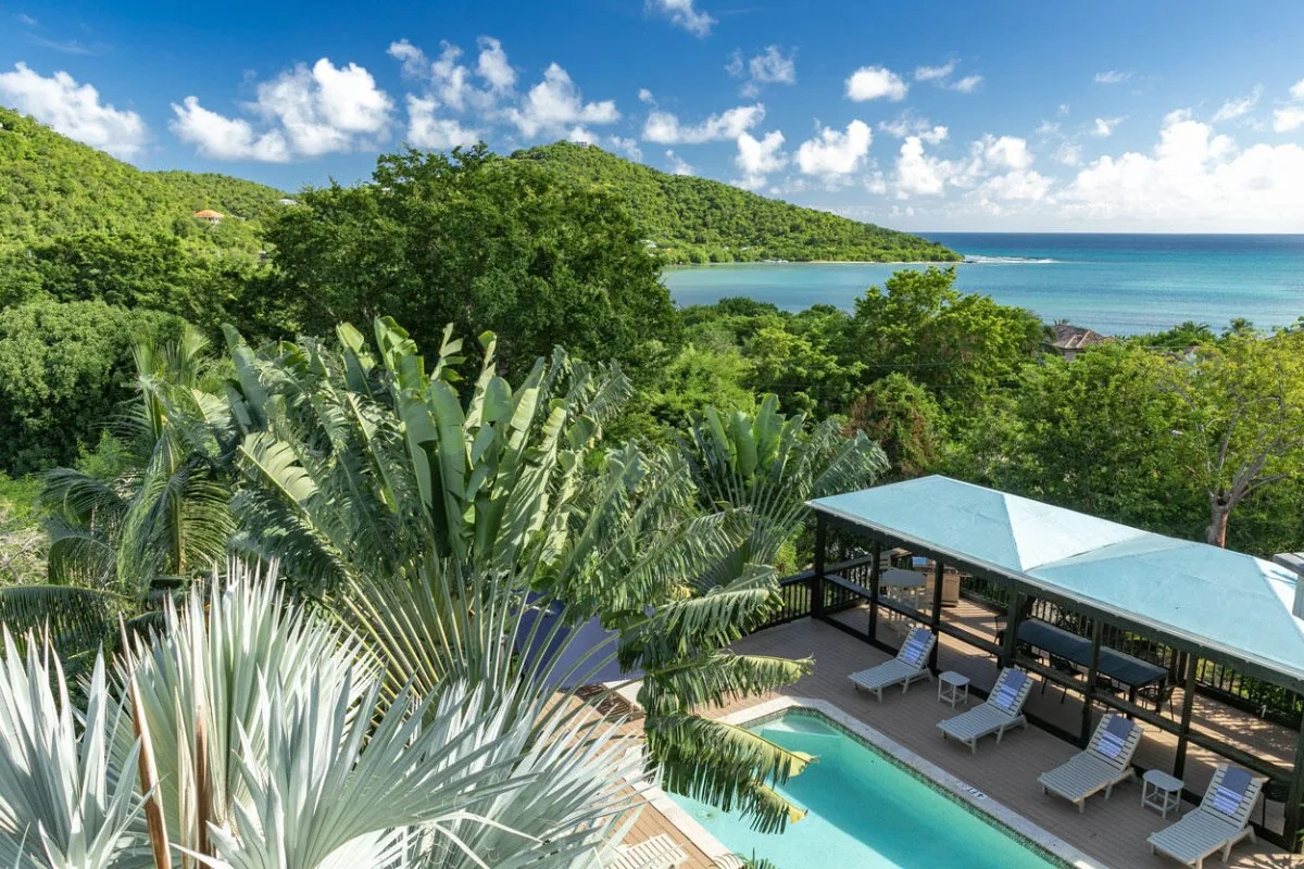 Una piscina tropical al aire libre junto a una zona cubierta con asientos y el océano al fondo.