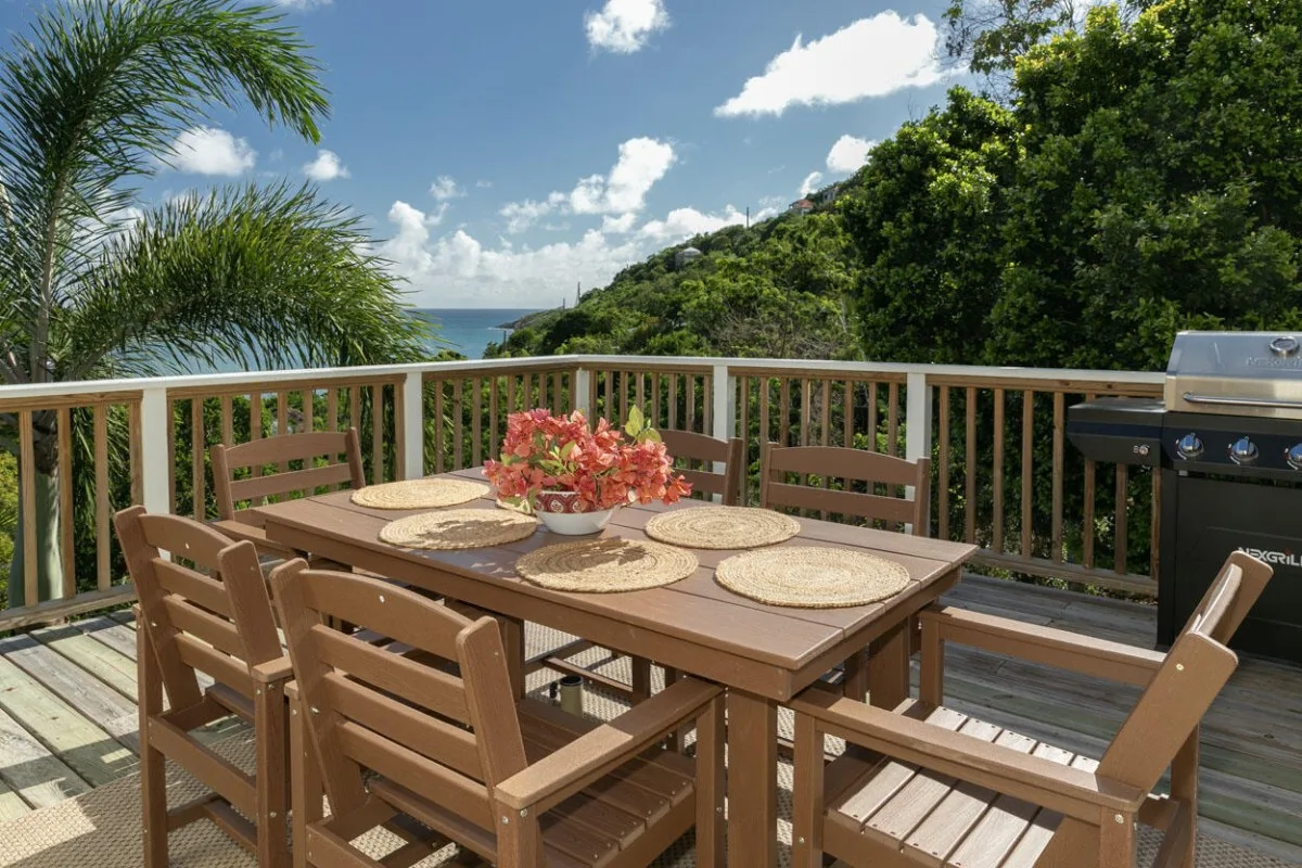 Una terraza comedor al aire libre con mesa de madera, asientos y parrilla, frente a vegetación y al mar.
