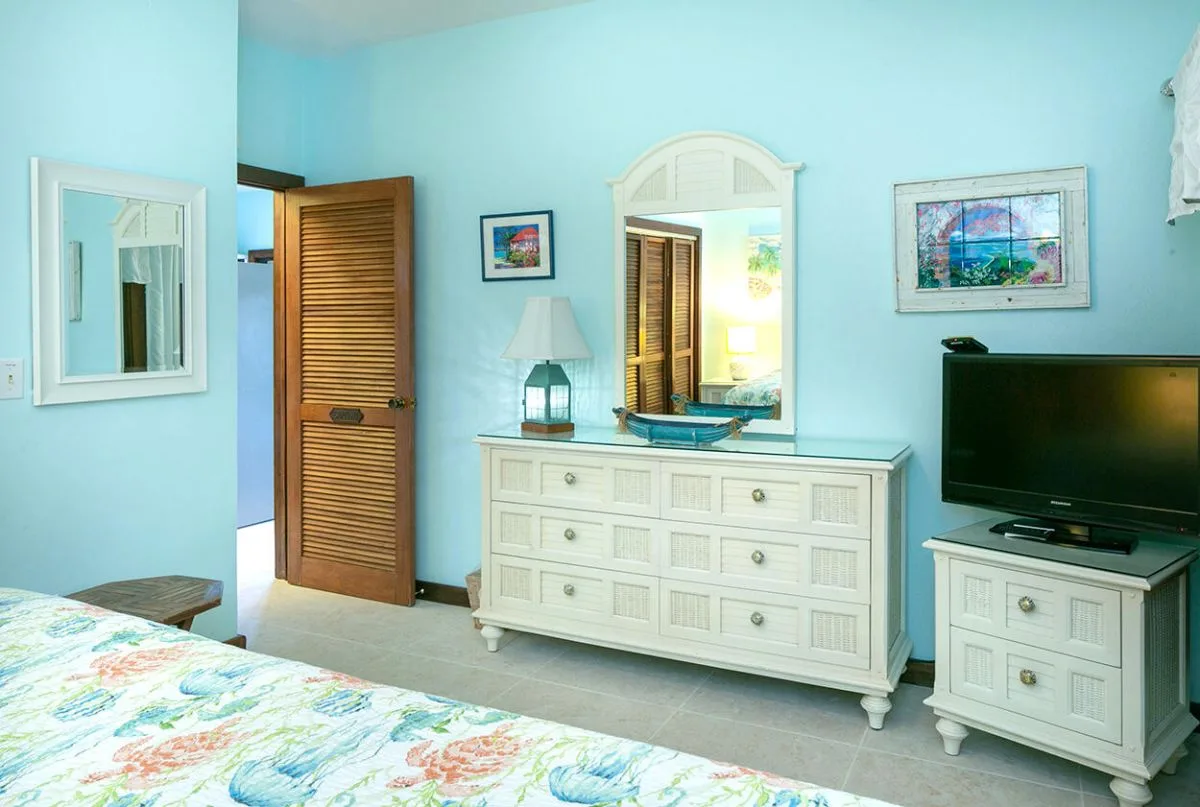 Un dormitorio luminoso con una cómoda blanca, un espejo y un televisor.