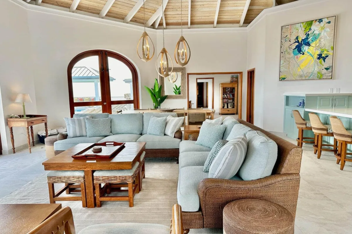A bright living room with cushioned seating, wooden tables, and exposed ceiling beams.