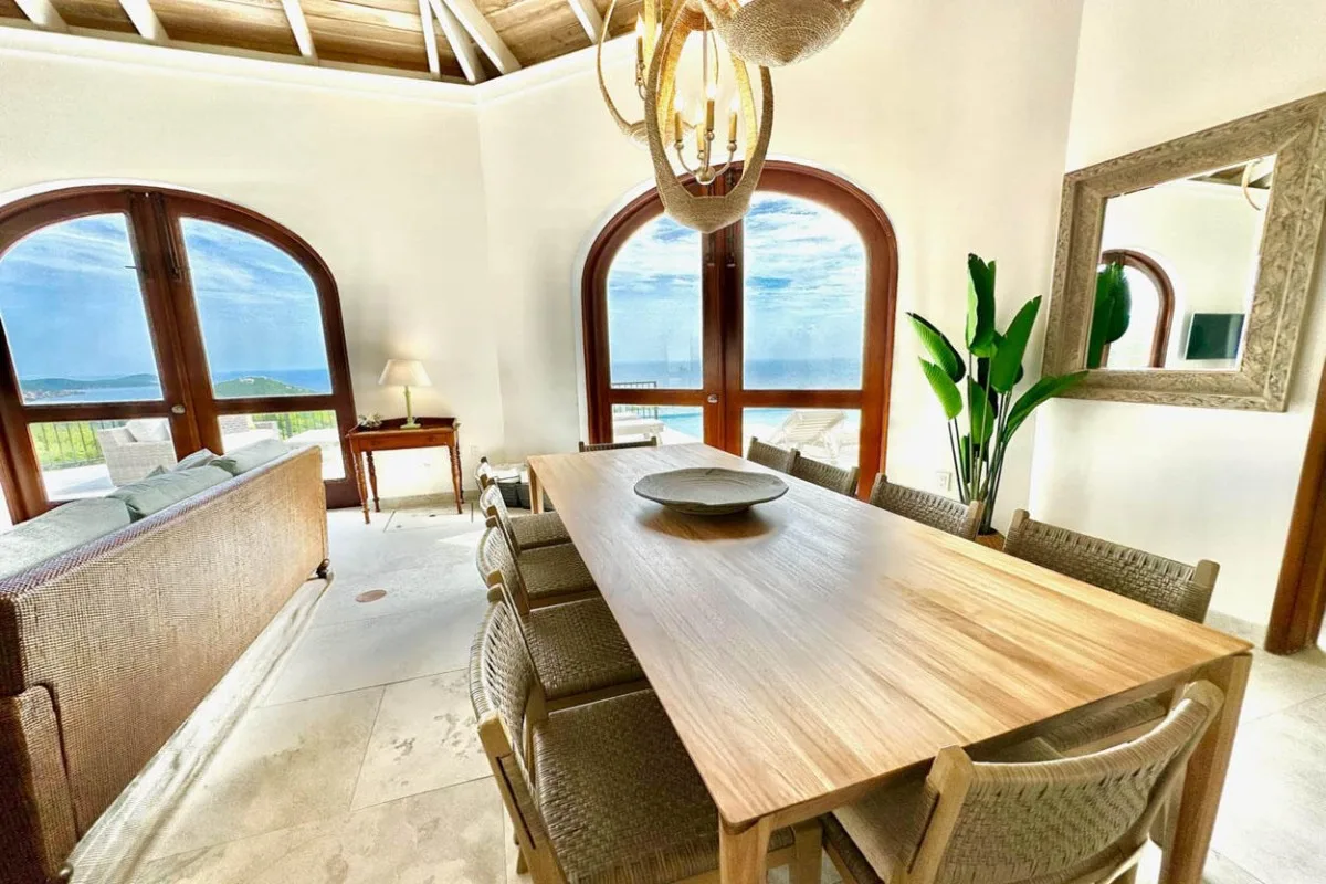 A bright dining area with a long wooden table beside arched windows.