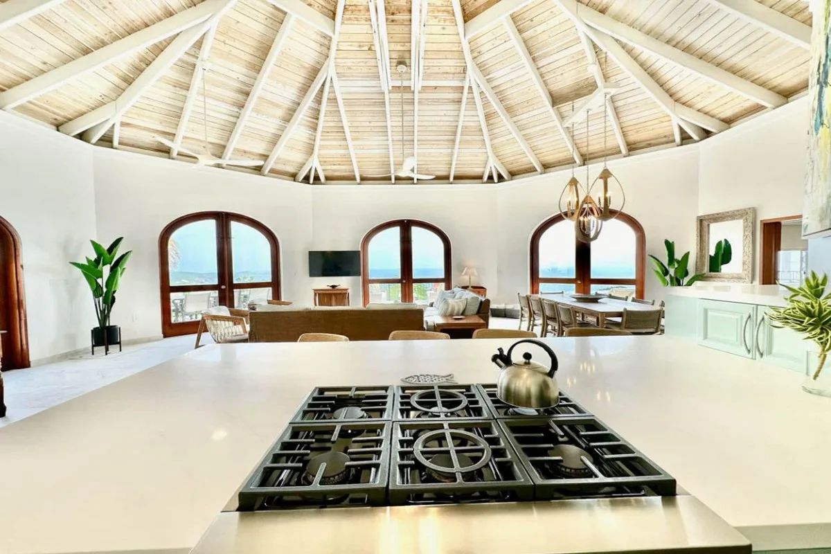 A bright kitchen with a large stovetop island beneath a high, exposed-beam ceiling.