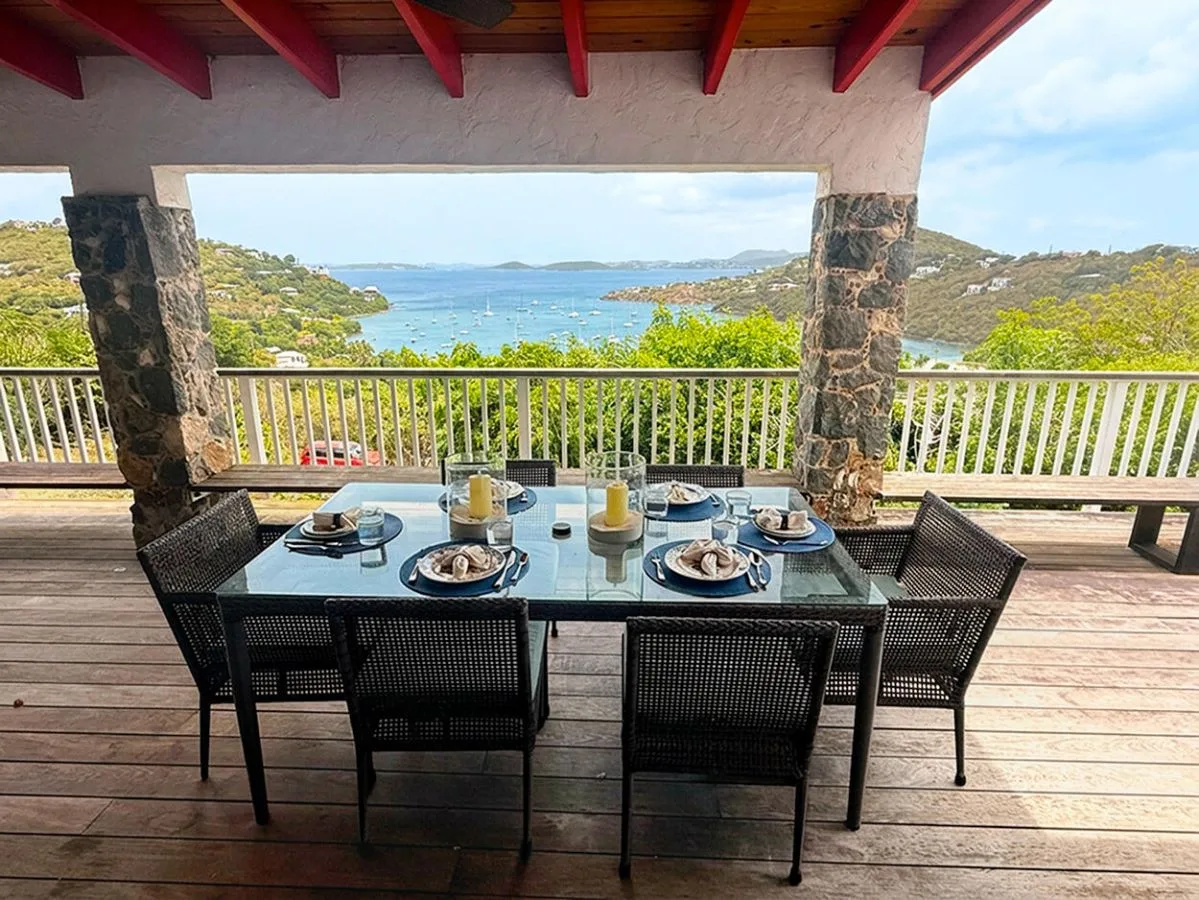 A covered outdoor dining area overlooks the water, with a set table and surrounding hills.