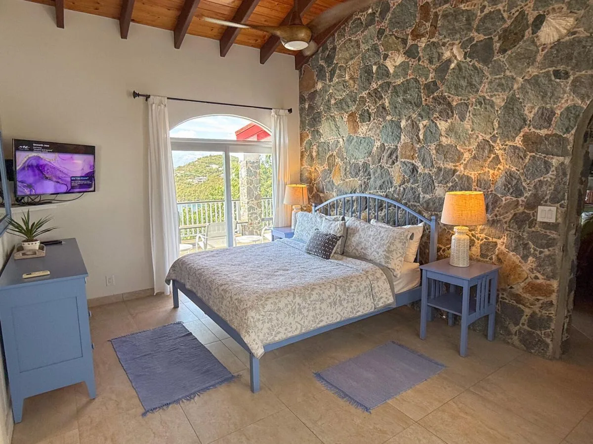 A bedroom with a bed beside a stone wall and a bright balcony doorway.