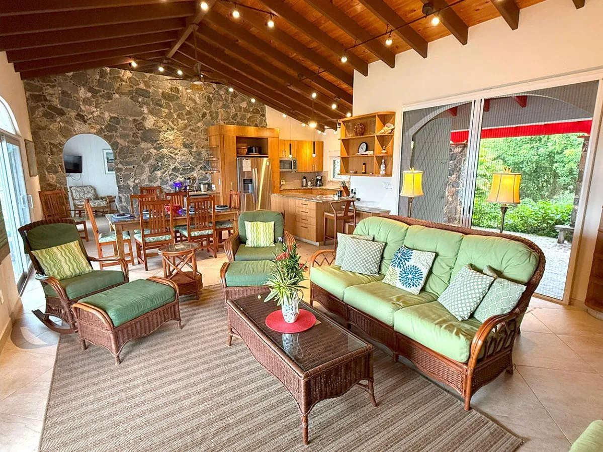 A living room with wicker seating and a dining area beneath a vaulted wooden ceiling.