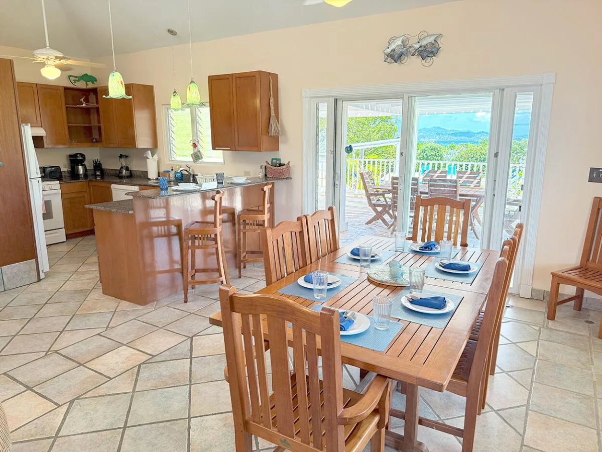 A dining area with wooden tables and chairs beside an open kitchen and large windows.