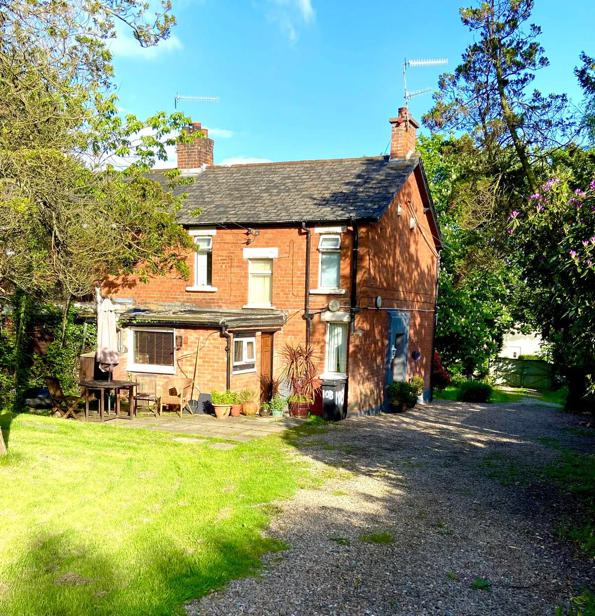 A red-brick house stands beside a gravel driveway, surrounded by trees and lawn.