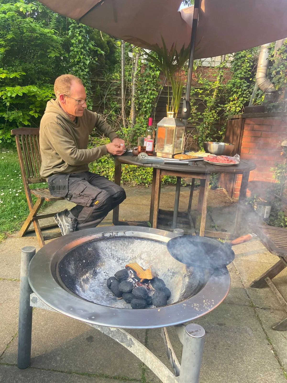 A garden patio with a man seated beside a smoking outdoor fire pit.