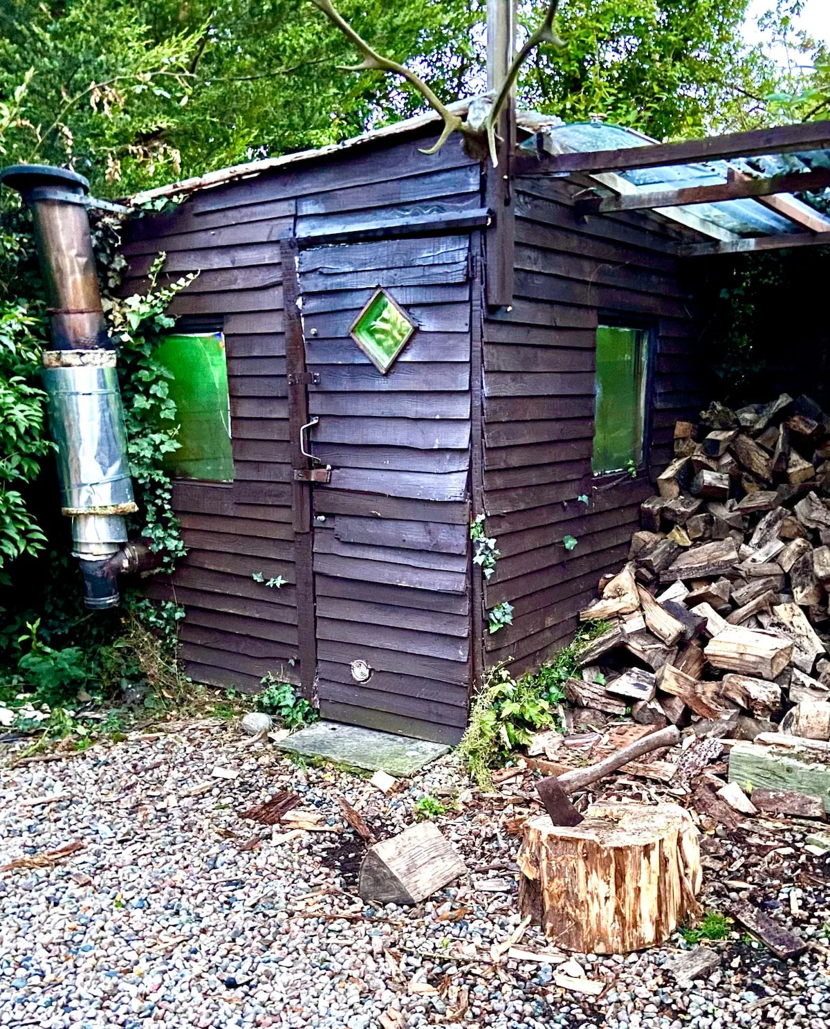 A small wooden outbuilding stands among trees beside a pile of chopped logs.